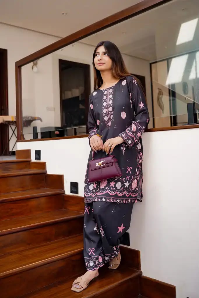 A woman wearing a dark purple and pink printed kurta set with matching pants, holding a purple handbag while standing on wooden stairs in a modern indoor setting.