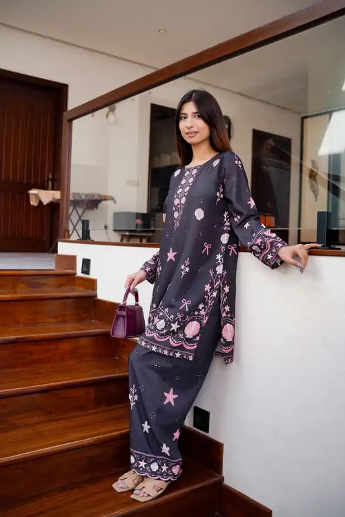 A woman stands on wooden stairs wearing a dark gray two-piece kurta set with pink and white celestial prints, holding a small purple handbag.